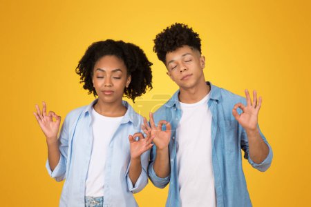 Téléchargez les photos : Femme afro-américaine détendue et homme les yeux fermés, méditant ou pratiquant la pleine conscience, faisant des signes OK avec leurs mains sur un fond jaune vif, incarnant la paix et la sérénité - en image libre de droit
