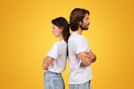 Photo for A couple in white t-shirts and blue jeans stands back to back with crossed arms and serious expressions, suggesting a disagreement or standoff, against a yellow background - Royalty Free Image