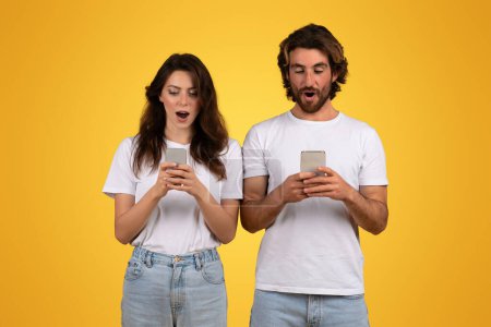 Photo for Astonished young man and woman in white t-shirts and blue jeans stand side by side, each engrossed and surprised by content on their smartphones against a yellow backdrop - Royalty Free Image