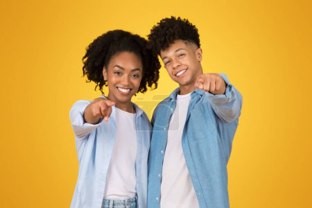 Téléchargez les photos : Confiant heureux Afro-Américain femme et homme pointant vers la caméra avec lumineux, sourires engageants, portant des vêtements décontractés sur un fond jaune monochrome - en image libre de droit