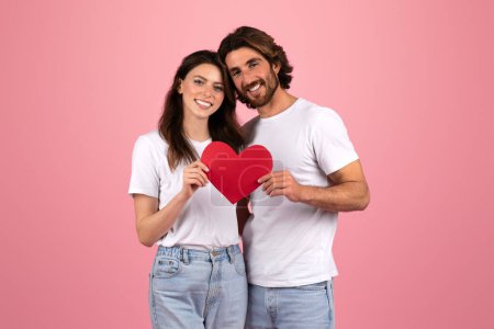 Téléchargez les photos : Heureux couple européen en t-shirts blancs et en jeans tenant ensemble un cœur en papier rouge, se regardant avec affection, symbolisant l'amour et la convivialité sur un fond rose - en image libre de droit