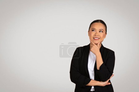 Téléchargez les photos : Femme d'affaires japonaise millénaire contemplative avec les bras croisés regarde vers le haut, son doigt sur son menton, dans un moment de réflexion réfléchie, sur un fond gris clair - en image libre de droit
