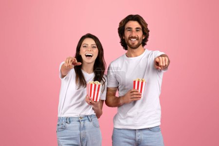 Téléchargez les photos : Joyeux couple européen en t-shirts et jeans blancs, riant et pointant vers la caméra, chacun tenant une petite boîte de pop-corn rayé, sur un fond rose doux, studio - en image libre de droit