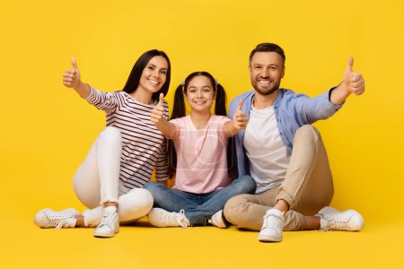 Photo for We Recommend. Portrait Of Happy Caucasian Family Of Three Showing Thumbs Up At Camera, Joyful Young Parents And Cute Daughter Gesturing Sign Of Approval, Posing On Yellow Background, Copy Space - Royalty Free Image