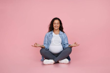 Téléchargez les photos : Méditation de grossesse, soulagement du stress. Jeune femme enceinte souriante faisant du yoga assis dans la pose de lotus sur le sol sur fond rose. Studio Shot - en image libre de droit