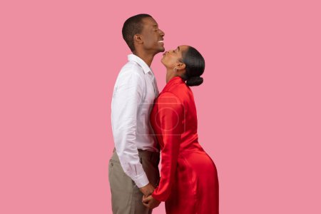 Foto de Mujer negra cariñosa en vestido rojo se inclina a besar al hombre sonriente en camisa blanca, tanto en pose amorosa sobre fondo rosa suave, vista lateral - Imagen libre de derechos