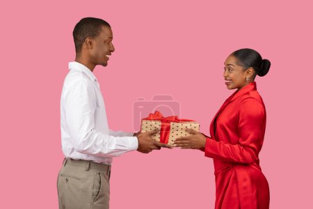 Photo for Side profile of cheerful black man handing wrapped gift with red ribbon to pleased woman in red dress, both showing happy expressions - Royalty Free Image