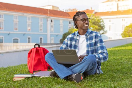 Téléchargez les photos : Sourire mec noir assis avec un ordinateur portable sur la pelouse verte dans le parc, websurf tout en apprenant et en se préparant à l'examen académique en dehors du campus universitaire, posant avec l'ordinateur regardant loin - en image libre de droit