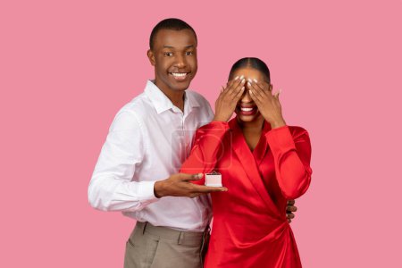 Photo for Happy black man presenting an engagement ring to an ecstatic woman with her hands over her eyes, moment of surprise and joy, pink background - Royalty Free Image
