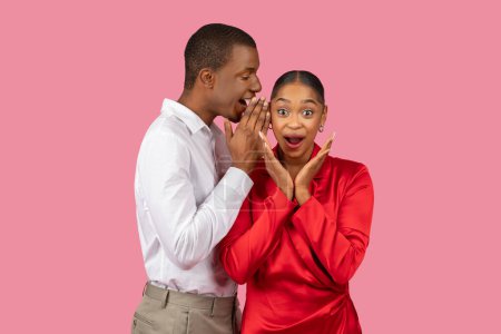 Photo for Black man in white shirt whispering into womans ear, her expression of delighted shock, hands on face, against playful pink background - Royalty Free Image