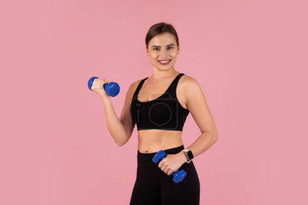 Photo for Energetic smiling woman exercising with blue dumbbells against pink studio background, sporty fit female wearing black athleticwear and smartwatch illustrating dynamic fitness session, copy space - Royalty Free Image