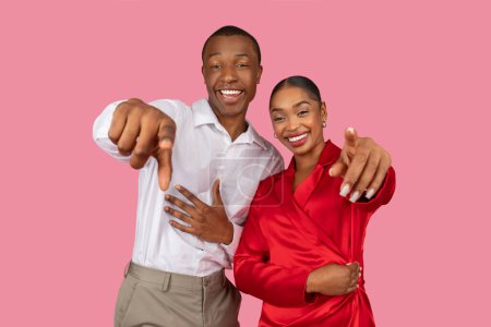 Photo for Smiling african american man and woman in formal attire pointing directly at camera, making cheerful and inviting gesture, standing against pink backdrop - Royalty Free Image