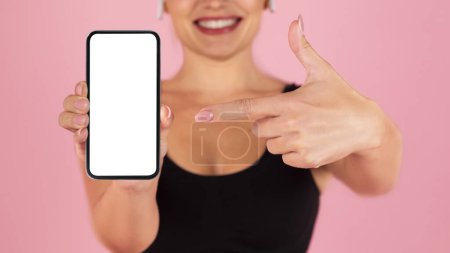 Photo for Cheerful young woman pointing to blank smartphone with white screen, selective focus on the phone, smiling unrecognizable female advertising new app, standing on soft pink studio background, mockup - Royalty Free Image