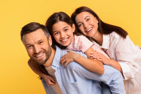 Téléchargez les photos : Portrait de la famille européenne joyeuse des parents et de la fille enfant étreignant ensemble, papa et maman posant embrassant leur enfant mignon sur fond de studio jaune, souriant à la caméra - en image libre de droit