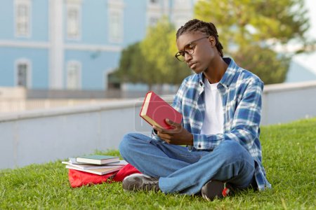 Téléchargez les photos : Un homme afro-américain réfléchi tenant un titre de lecture de livre, apprenant la littérature sur la pelouse verte, un étudiant lit un roman assis avec un sac à dos et des cahiers en plein air au parc du campus - en image libre de droit