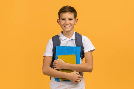 Foto de Escolar adolescente sonriente con mochila sosteniendo libros de texto y sonriendo a la cámara, niño varón feliz vestido de polo blanco de pie sobre fondo de estudio amarillo, retratando el entusiasmo por la educación - Imagen libre de derechos