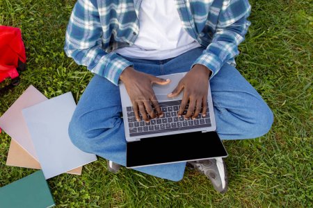 Téléchargez les photos : Plan recadré de l'homme afro-américain tapant sur ordinateur portable assis sur la pelouse verte, apprendre en ligne au parc en plein air, gars websurfing sur ordinateur combinant la technologie et les loisirs, plan grand angle - en image libre de droit