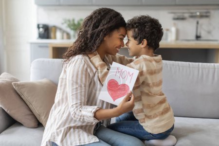 Photo for Adorable black little son giving handmade card with drawn heart to his mother at home, loving mom and male child having moment of love and appreciation, relaxing in comfort of their living room - Royalty Free Image