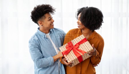 Téléchargez les photos : Un jeune couple afro-américain joyeux partage un moment spécial, échangeant un cadeau joliment emballé avec un ruban rouge, leurs visages illuminés de sourires dans une pièce légère et aérée - en image libre de droit