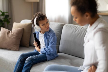 Téléchargez les photos : Jeune garçon noir portant des écouteurs montre expression mécontente tandis que sa mère semble aborder le problème sur le canapé dans leur maison - en image libre de droit