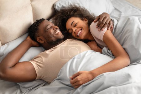 Téléchargez les photos : Aimant jeune homme et femme noirs allongés dans leur lit embrassant le bonheur rayonnant, leur étreinte décontractée témoigne de leur relation insouciante et aimante, profitant d'une matinée confortable ensemble dans la chambre - en image libre de droit