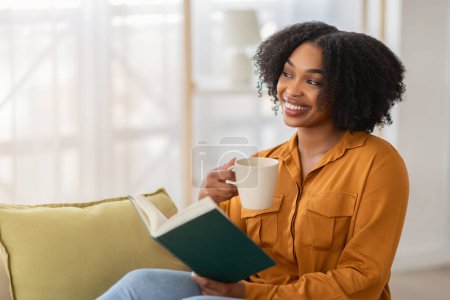 Photo for Cheerful young african american lady enjoy cup of coffee, spare time, rest and read book, sit on sofa in living room interior. Relax, study with favorite drink, hobby at home - Royalty Free Image