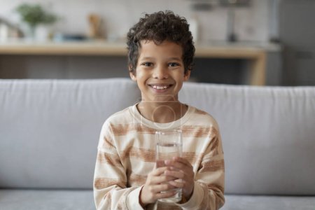 Photo for Cheerful little black boy with bright smile holding clear glass of water, smiling african american male child sitting on sofa in well-lit living room, promoting healthy hydration habits, copy space - Royalty Free Image