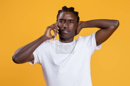 Photo for Annoyed young black man talking on cellphone and touching his head, confused african american guy having mobile call, displaying frustration during phone conversation, standing on yellow background - Royalty Free Image