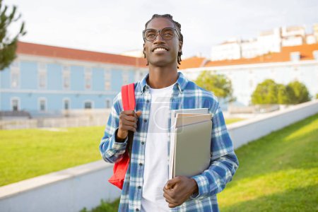 Foto de Hombre afroamericano estudiante en gafas posa con el ordenador portátil, mochila y sus libros de trabajo, de pie cerca del edificio de la universidad al aire libre, sonriendo a la cámara con confianza. Alegría de graduación - Imagen libre de derechos