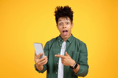 Photo for Emotional handsome stylish millennial black guy pointing at phone in his hand and exclaiming, looking at camera, isolated on yellow studio background. Online offer, deal, promo concept - Royalty Free Image