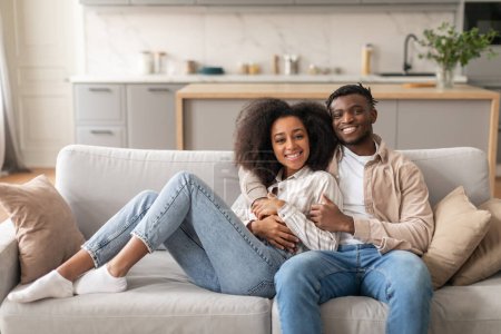 Téléchargez les photos : Heureux couple afro-américain romantique étreignant tout en se refroidissant sur le canapé à l'intérieur de la maison. Jeunes conjoints jouissant du repos et de la détente le week-end, assis ensemble dans le salon - en image libre de droit