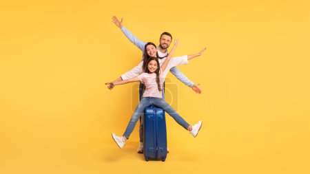 Téléchargez les photos : Vacances en famille. Joyeux parents européens et petite fille posant avec une valise de voyage, écartant les bras comme un avion, prêts pour un voyage en avion ensemble, sur fond de studio jaune. Panorama, Espace de copie - en image libre de droit