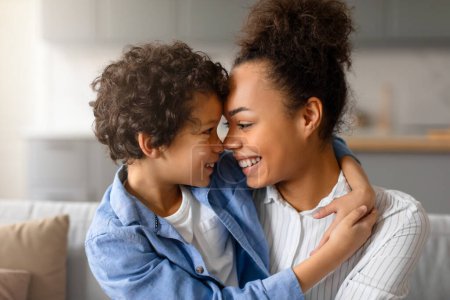 Téléchargez les photos : Délicieuse mère noire et son fils touchant affectueusement les fronts, partageant un moment heureux et intime dans leur salon confortable et ensoleillé à la maison - en image libre de droit