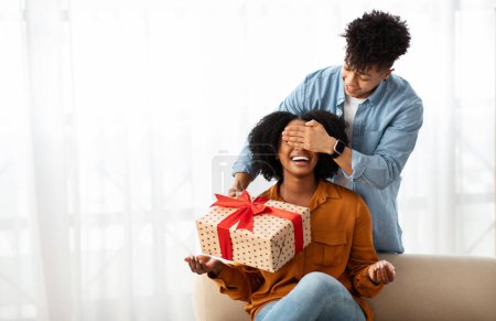 Téléchargez les photos : Souriant jeune homme afro-américain les yeux fermés à dame, donne boîte-cadeau, dans le salon intérieur. Surprise, amour et relation, anniversaire, fête à la maison - en image libre de droit