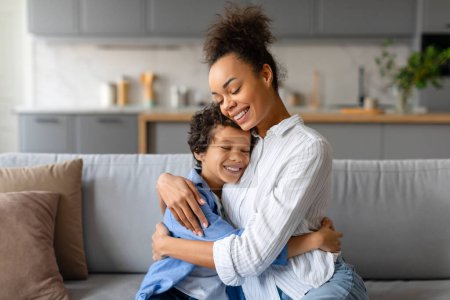 Photo for Joyful african american mother embraces her little son with loving hug, sharing moment of affection in their bright, modern kitchen interior - Royalty Free Image