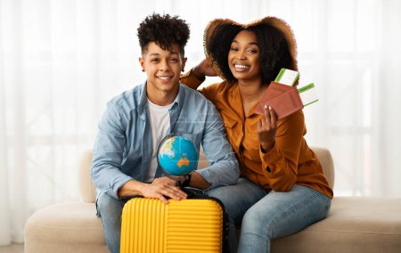 Téléchargez les photos : Souriant jeune couple afro-américain en chapeau, avec valise, passeport de spectacle et billets dans l'intérieur du salon. Aventure de vacances ensemble, relation, amour, voyage romantique, voyage - en image libre de droit