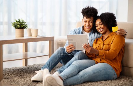 Photo for Happy young african american guy and woman shopaholics, hugs, use tablet, credit card, enjoy pay for order in living room interior. App, sale, shopping at free time at home - Royalty Free Image