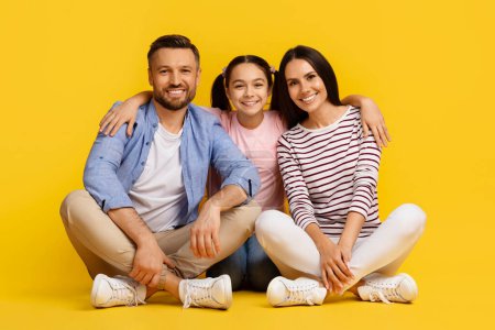 Téléchargez les photos : Famille caucasienne avec une fille adolescente assise sur le sol et souriant vers la caméra, de jeunes parents heureux et leur enfant femelle embrassant et collant, posant isolé sur fond jaune - en image libre de droit