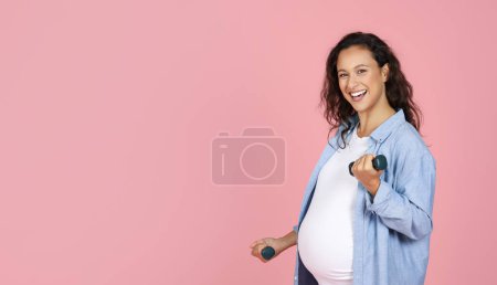 Téléchargez les photos : Femme enceinte souriante faisant de l'exercice avec des cloches, regardant la caméra et souriant, panorama avec espace de copie. Femme enceinte saine formation sur fond rose, à l'aide d'haltères. Sport pendant la grossesse - en image libre de droit
