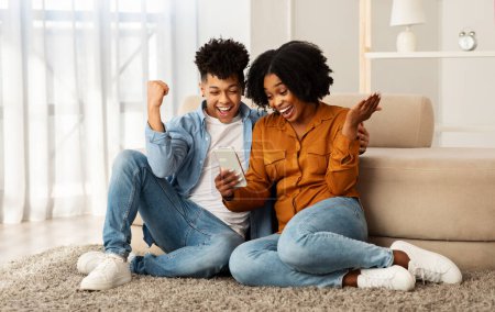 Photo for Glad excited young black couple winners, with smartphone celebrate win, success, rise fist, sit on floor, good news in living room interior. App, video call, ad and offer, emotions - Royalty Free Image