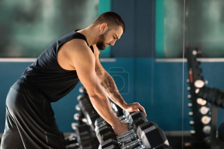 Photo for Sport Equipment Concept. Young Male Athlete Taking Dumbbell From Rack At Gym, Portrait Of Masculine Tattooed Man Choosing Barbells, Preparing For Bodybuilding Workout At Modern Sport Club, Side View - Royalty Free Image