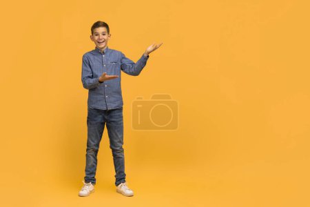 Téléchargez les photos : Adolescent joyeux garçon en faisant un geste de présentation avec ses mains, adolescent souriant garçon debout sur fond de studio jaune, parfait pour le placement de produits ou de démontrer des caractéristiques, copier l'espace - en image libre de droit