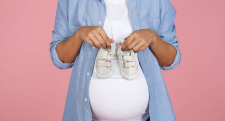 Photo for Cropped of pregnant woman holding cute white little boots for unborn baby on her big belly, waiting for child, isolated on pink studio background. Pregnancy, motherhood concept - Royalty Free Image