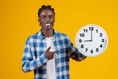Foto de Joven hombre negro apuntando a un gran reloj de pared blanco en su mano y sonriendo a la cámara, alegre hombre afroamericano que representa la gestión del tiempo y la puntualidad, de pie sobre el fondo amarillo - Imagen libre de derechos
