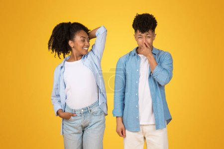 Photo for A cheerful young African American woman confidently posing with a hand on her hip, while the young man beside her laughs and covers his nose, against a vivid yellow background - Royalty Free Image