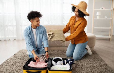 Photo for A smiling african american couple is packing for a trip, with the woman handing clothes to the man who is organizing the suitcase, both surrounded by a cozy home environment - Royalty Free Image