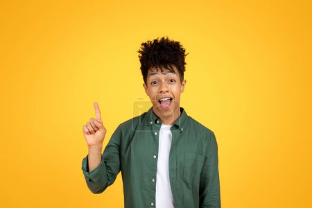 Foto de Emocionado joven afroamericano chico con estilo peinado levantando el dedo hacia arriba y mueca, tienen buena idea creativa o gran solución, fondo de estudio amarillo - Imagen libre de derechos