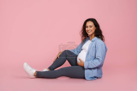 Photo for Pregnancy, waiting for baby. Happy attractive pregnant young woman wearing casual clothing sitting on floor isolated on pink studio background, touching her big tummy, copy space - Royalty Free Image