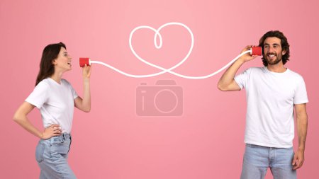 Foto de Una joven pareja europea se comunica juguetonamente a través de la lata de lata de teléfonos conectados por una cadena que forma una forma de corazón, simbolizando el amor y la conexión sobre un fondo rosa - Imagen libre de derechos