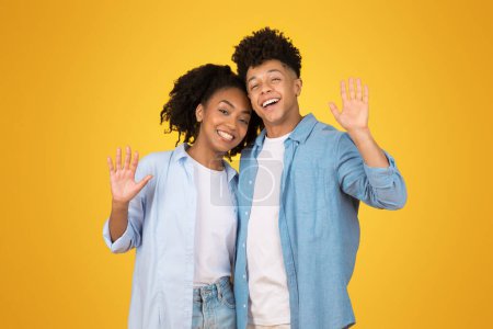 Téléchargez les photos : Affable jeune femme afro-américaine heureuse et homme agitant bonjour avec les mains ouvertes, à la fois des sourires brillants sportifs et tenue bleue décontractée, dans un cadre jaune joyeux - en image libre de droit